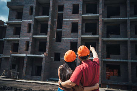 Back view a man and a woman in orange helmets stand with their arms around each other and look at a brick apartment building under construction. Investment in apartment, mortgage construction.の写真素材