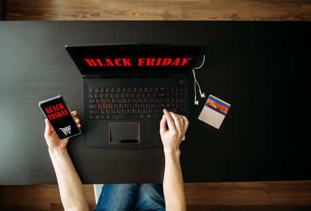 Inscription Black Friday. Online Shopping Concept. Cropped view woman's hand holding smartphone with app and using laptop computer.の写真素材