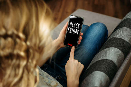 Black Friday shopping app in a mobile phone screen. Woman holding smartphone in the hand, lying on sofa. Safety shopping from home during the quarantine.の写真素材