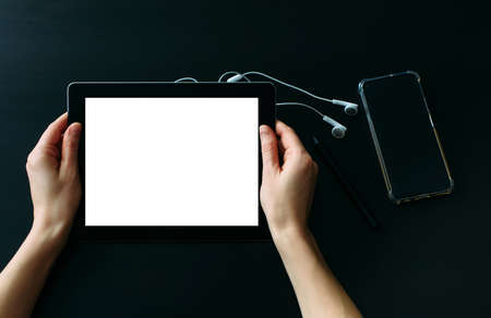 Women's hands holding tablet computer with empty white screen against black wooden table, on which...