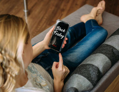 Black Friday shopping app in a mobile phone screen. Woman holding smartphone in the hand, lying on sofa. Safety shopping from home during the quarantine.の写真素材