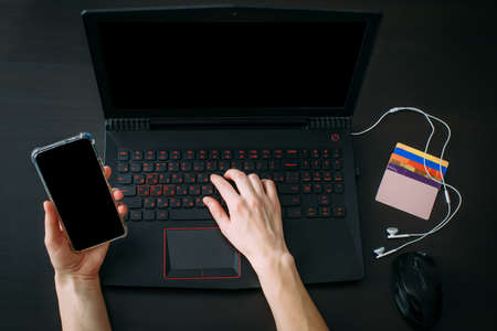 Top view woman's hand holding smartphone, laptop computer with blank screen on black background. Using gadgets for business, searching information in workplace at home office.の写真素材