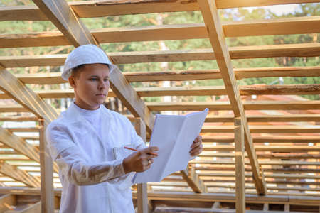 Foreman holding drawings, looking the project of wooden house, working on a construction site. Young attractive engineer in a white helmet checks the roof frame. Concept career architect. Copy space.の写真素材