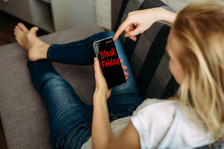 Black Friday shopping app in a mobile phone screen. Woman holding smartphone in the hand, lying on sofa. Safety shopping from home during the quarantine.の写真素材