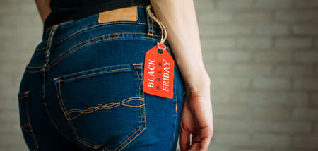 People, sale, consumerism concept. Close up cropped photo of sexual woman's buttocks wearing blue casual denim pants on gray brick background in the mall. Red tag black Friday on jeans.の写真素材