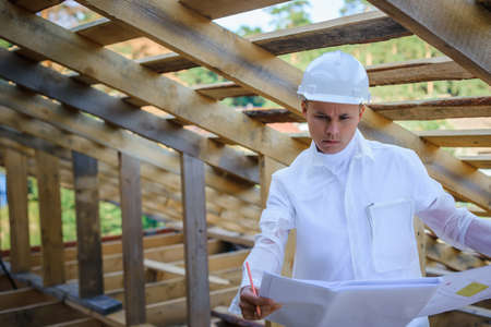 Foreman holding drawings, looking the project of wooden house, working on a construction site. Young attractive engineer in a white helmet checks the roof frame. Concept career architect. Copy space.の写真素材