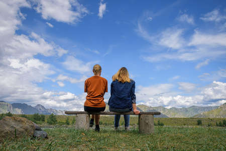 Two young women sitting on wooden bench, backs to the camera against beautiful landscape. Friends travel, admire magnificent view of mountain range under blue sky on sunny day.の写真素材