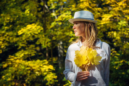 Beautiful elegant woman in white shirt and hat posing with bouquet of autumn leaves in sunny day. Young blonde holds bright yellow maple leaves in her hands. Natural blurred background, copy space.の写真素材