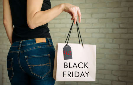 Cropped view sexy woman in denim holding paper bag with tag in her hand against a white brick wall in the mall. Copy space. Black Friday sale concept.の写真素材