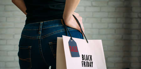 Woman holding a paper bag with tag black Friday. Back view of slender female butt in blue jeans. Concept of shopping, seasonal discounts.の写真素材