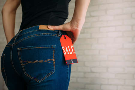 People, sale, consumerism concept. Close up cropped photo of sexual woman's buttocks wearing blue casual denim pants on gray brick background in the mall. Red tag black Friday on jeans.の写真素材