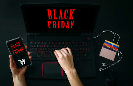 Inscription Black Friday. Online Shopping Concept. Cropped view woman's hand holding smartphone with app and using laptop computer.の写真素材