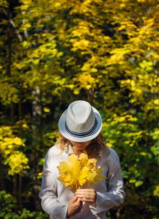 Beautiful young woman in white clothes and hat tilted her head to the bouquet of yellow leaves in her hands. Elegant blonde, face hidden by hat. Freedom, love, happiness. Indian summer, golden autumn.の写真素材