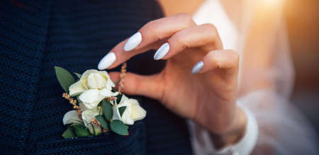 Bride's hand touches the buttonhole on the groom's jacket, close-up. Stylish suit, boutonniere of fresh flowers, woman's hand with a manicure.の写真素材