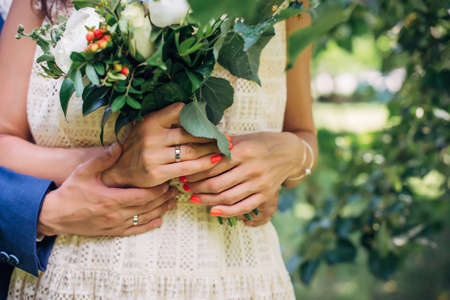 Hands of the bride and groom with wedding rings, bouquet of fresh flowers, vintage lace dress. Husband hugs his wife. Concept of marriage, love, and family life.の写真素材