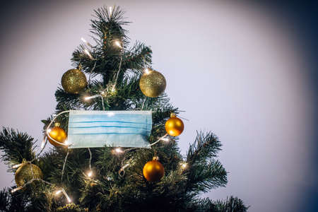 Medical protective mask hanging on Christmas tree decorated with shiny balls and glowing garland. Celebrating winter holidays safely during coronavirus pandemic. Covid and happy new year concept.の写真素材