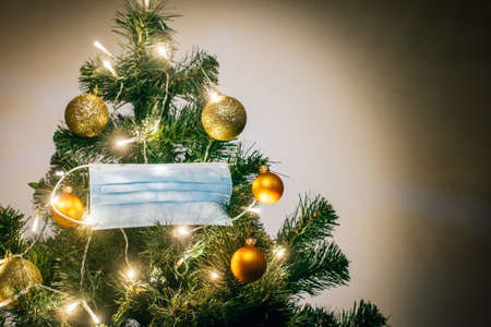 Medical protective mask hanging on Christmas tree decorated with shiny balls and glowing garland. Celebrating winter holidays safely during coronavirus pandemic. Covid and happy new year concept.の写真素材