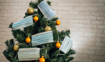 Christmas tree decorated with blue medical masks and golden balls on the background of light wall in apartment. Concept of the new year 2021, coronavirus pandemic. Holiday in quarantine. New normal.の写真素材