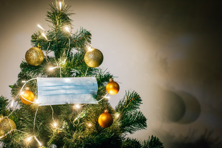 Medical protective mask hanging on Christmas tree decorated with shiny balls and glowing garland. Celebrating winter holidays safely during coronavirus pandemic. Covid and happy new year concept.の写真素材