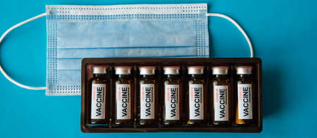 Box of ampoules with label and medical mask on blue background with copy space, close-up. Coronavirus vaccine ready for use. Mass vaccination of the population.の写真素材