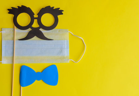 Medical face mask and paper glasses on stick on bright yellow background, close-up. April Fools' Day and coronavirus pandemic. Accessories for jokes and parties.の写真素材