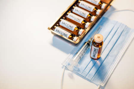 Concept of vaccination against coronavirus. Bottle of vaccine on blue medical mask, syringe and box of ampoules, white background, selective focus. Lettering on the label Covid-19. Copy space.の写真素材