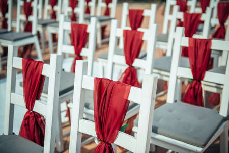 White wooden chairs decorated with red fabric and ribbons for wedding registration outdoor. Guest chairs in rows, close up.の写真素材