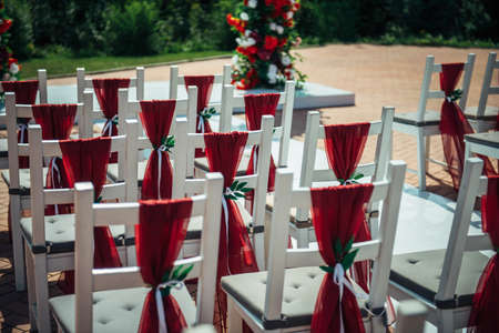 White wooden chairs decorated with red fabric and ribbons for wedding reception outdoor. Guest chairs in rows in the summer park, blurred background.の写真素材