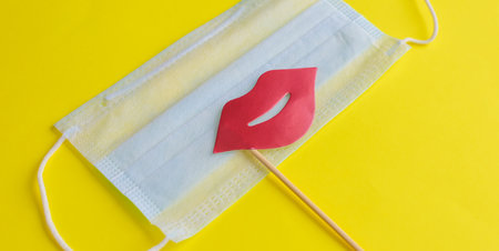 Medical face mask, paper pink lips on stick on bright yellow background, close-up. Celebrating April Fools' Day during coronavirus pandemic concept.の写真素材