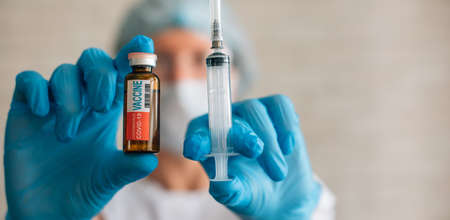 Female doctor or nurse in uniform and blue gloves holds vial and syringe. Focus on label text: COVID-19 VACCINE. Coronavirus immunization concept.の写真素材