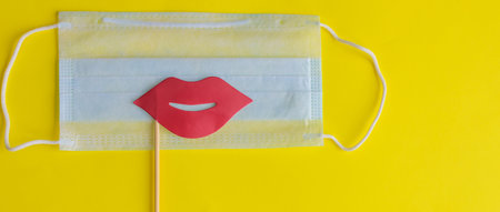 Medical face mask, paper pink lips on stick on bright yellow background, close-up. Celebrating April Fools' Day during coronavirus pandemic concept.の写真素材