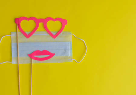 Medical face mask, paper glasses and pink lips on stick on bright yellow background, close-up. April Fools' Day during coronavirus pandemic concept. Copy space.の写真素材