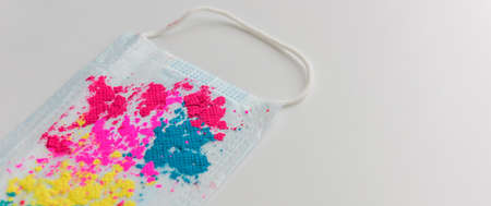 Blue medical face mask, with bright multicolored holi paints, on white background, close-up. Concept of celebrating indian spring festival of colors during covid-19 pandemic. Copy space.の写真素材
