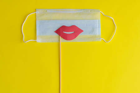 Medical face mask, paper pink lips on stick on bright yellow background, close-up. Celebrating April Fools' Day during coronavirus pandemic concept.の写真素材