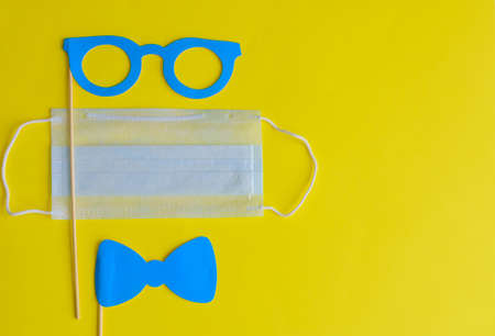 Medical face mask and paper glasses on stick on bright yellow background, close-up. April Fools' Day and coronavirus pandemic. Accessories for jokes and parties.の写真素材