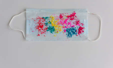 Blue medical mask, sprinkled with multicolored holi paints on white background, close-up. Concept of celebrating the spring festival of colors during coronavirus pandemic. Copy space.の写真素材
