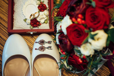 Vintage wedding jewelry and accessories, close-up. Gold wedding rings, ruby earrings, classic bride's shoes, bouquet of fresh red and white roses.の写真素材