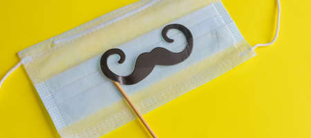 Medical face mask, paper black mustache on a stick on bright yellow background, close-up. Celebrating April Fools' Day during the coronavirus pandemic.の写真素材