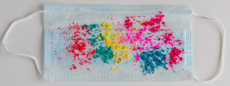 Blue medical mask, sprinkled with multicolored holi paints on white background, close-up. Concept of celebrating the spring festival of colors during coronavirus pandemic.の写真素材