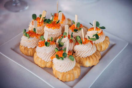 Plate of cold appetizers on white tablecloth, close-up. Profiteroles with cream cheese and red caviar filling. Delicacies at festive banquet.の写真素材