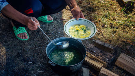 Hands of adult woman pouring soup from a cauldron into a plate with ladle. Cooking in the fresh air on a hiking trip. Large pot standing on the grass and firewood. Concept of tourism.の写真素材