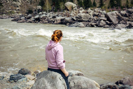 Lonely young brunette woman in sad mood sitting back to the camera on big gray stone near stormy...
