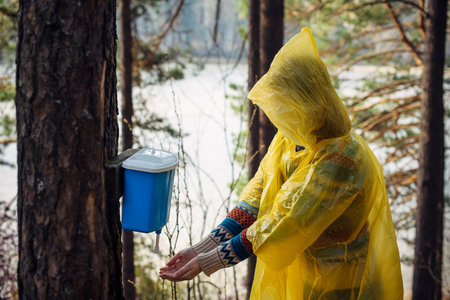 Unknown woman in yellow raincoat washes her hands in wash basin hanging on tree. Rainy morning on tourist camp in forest near the river.の写真素材