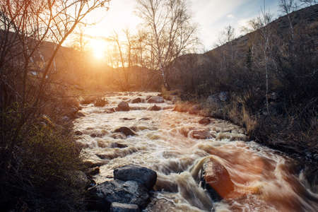 Small mountain river surrounded by rocky banks against the hills and sunset. Dry grass and leafless...