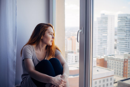 Young beautiful red-haired woman sits on the windowsill and looks at urban landscape outside the window. Portrait of a pensive long-haired girl in a city apartment.の写真素材