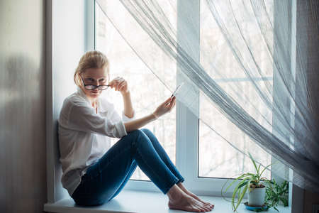 Barefoot woman in glasses and white shirt sitting on the windowsill, holding smartphone in her hand. Cute blonde looking at camera, close-up.の写真素材