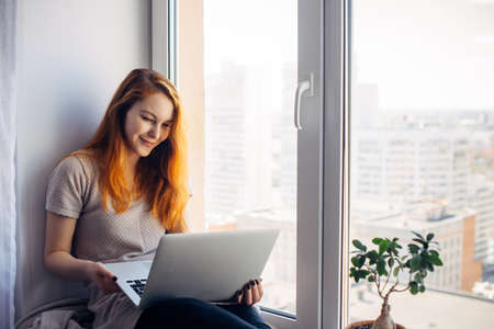 Young beautiful woman with long red hair sits on windowsill and looks at laptop. Female writer with...