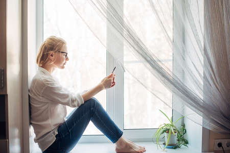 Pretty blonde woman with bare feet in glasses, jeans and white shirt sitting on the windowsill in...