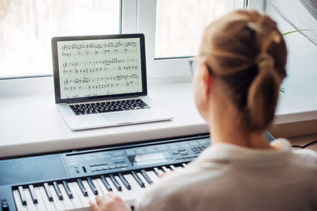Back view young woman playing a synthesizer, reading notes on a laptop screen, close up. Independent learning to play the piano at home. Passion for music, hobbies, leisure, self-development.の写真素材