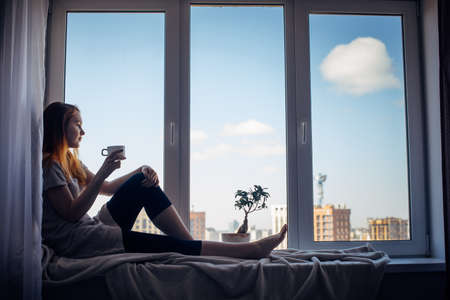 Silhouette of young slender girl sitting on the windowsill at home, side view, copy space. Outside the window blue sky and tall city buildings. Red-haired woman holding cup, looking at the metropolis.の写真素材